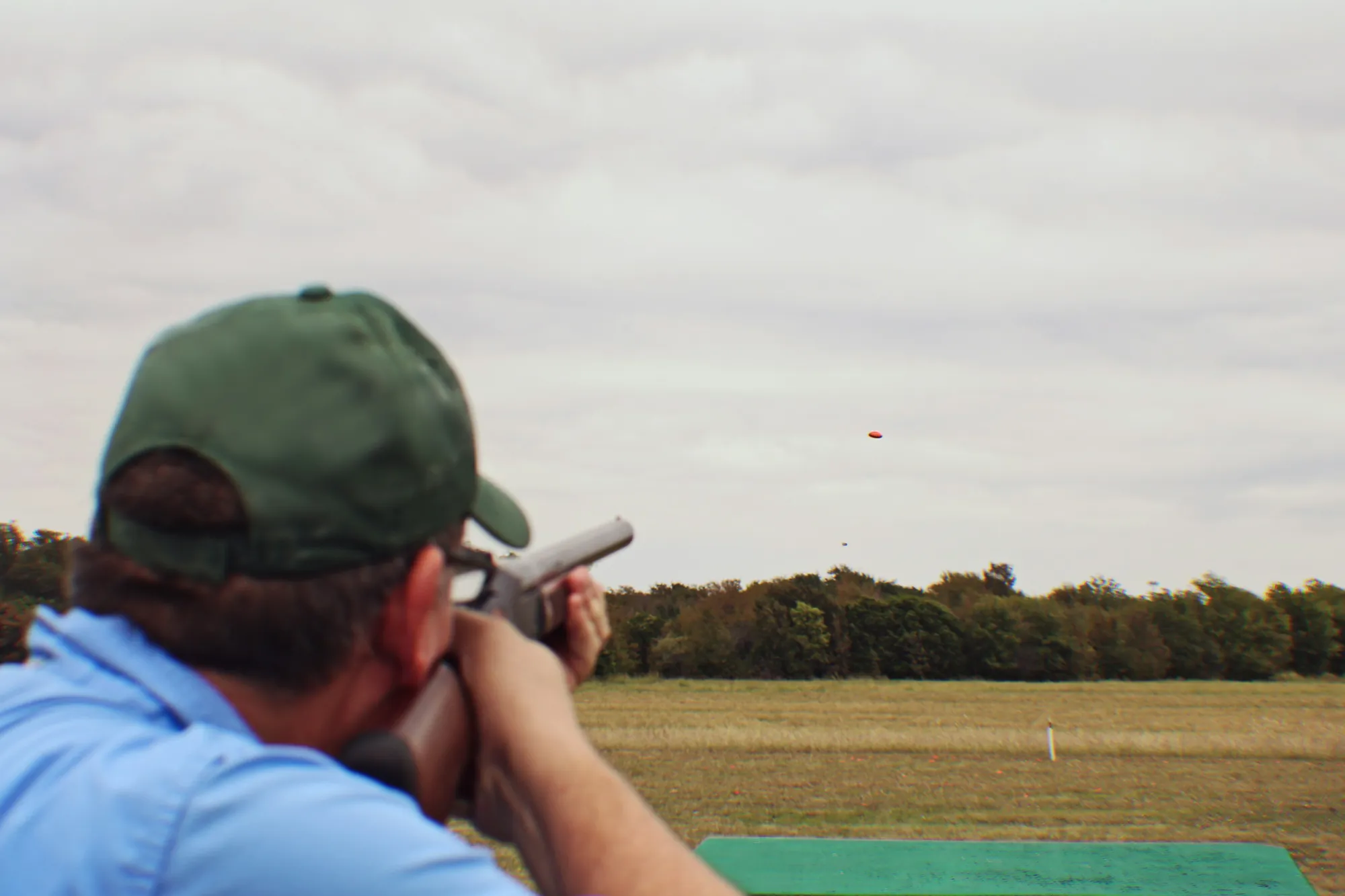 A shooter in a green cap on a clay ground, gun raised, a clay in the air against a wide grey sky